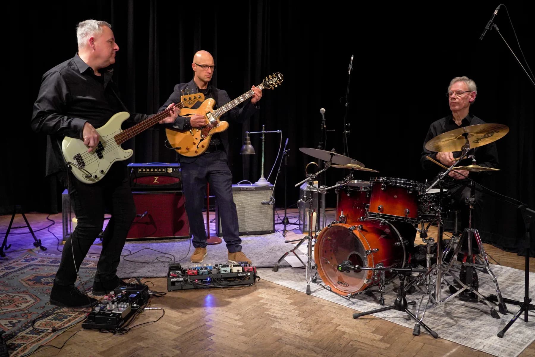 Pete Roth Trio performance photo with guitar and drums in dramatic stage light.